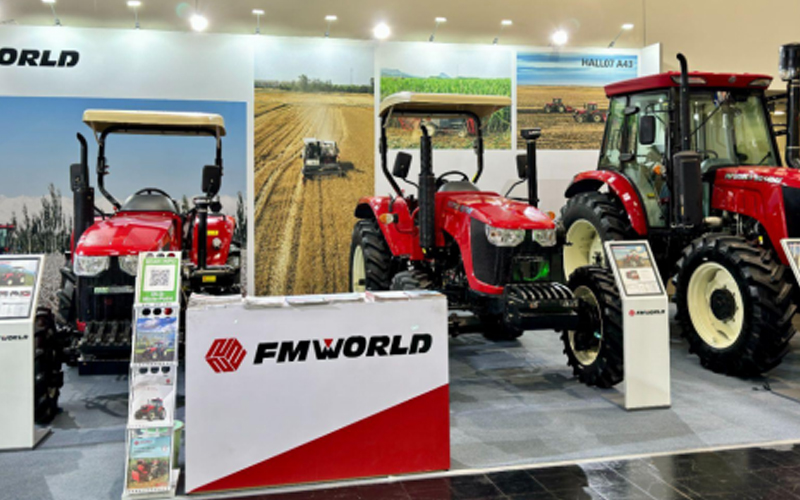 ထင်ရှားသော 'FMWORLD' အမှတ်တံဆိပ်ဖြင့် လယ်ယာမြေရှုခင်းများကြားတွင် ကျစ်လစ်သော၊ အလတ်စားနှင့် အရွယ်အစား-ကြီးမားသော လယ်ထွန်စက်သုံးစီးပါဝင်သော စိုက်ပျိုးရေးကုန်စည်ပြပွဲပြခန်း။
